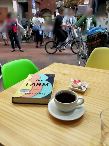 The photo is of a light wooden square table on which is a white mug & saucer of black coffee. To the left is the hardcover library book THE FARM with 3 colored silhouette profiles of pregnant torsos - yellow, red, & turquoise. Above the coffee is a tiny square white saucer with a plastic colored shrimp cracker and tiny chocolate. The right chair is green, the left one yellow. In the distance are blurred Japanese adults & children. A woman in a black one piece dress is pedaling to the right. There are adults protecting children from such traffic because it is festival time and the children are helping with activities, dressed in blue happi jackets. One man's happi is green. A policewoman is on the right in a red police jacket.