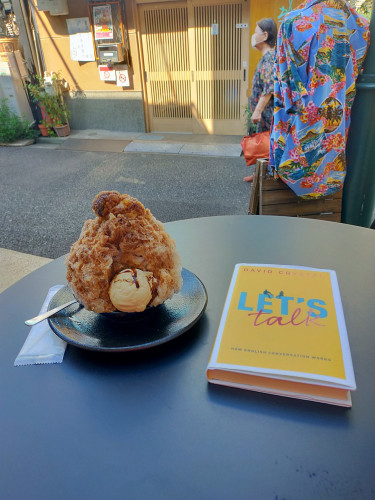 Photo is of a round black metal table outside a cafe. On it are the yellow hard back book with LET'S in bold turquoise TALK in lowercase cursive red. To the right is a brown mound of shaved ice with a scoop of vanilla ice cream in front on a black table. A slatted tan wooden sliding door can be seen in the distance. A Hawai'ian shirt can be seen hanging off a torso mannequin to the right and an old masked Japanese woman holding an orange purse can be seen emerging behind it