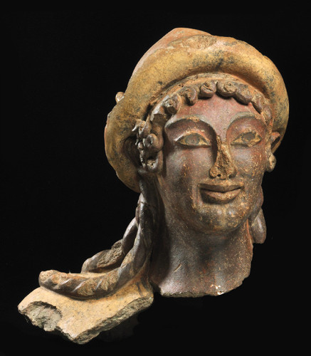 Etruscan terracotta head of the god Turms, associated with the Greek god Hermes. He is wearing a pilos hat, his hair arranged in neat curls and long braids falling to his shoulders. The god has large, expressive eyes and a kind smile.