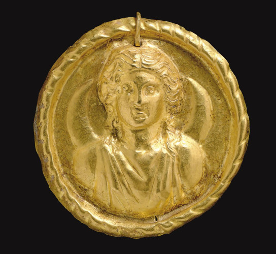 A Roman gold medallion of the moon goddess Luna. The sheet disk with a repoussé bust of the moon goddess depicted frontally wearing a sleeveless tunic, her center-parted hair rolled back along the sides of her face and falling along her neck, a crescent moon behind her, the medallion with a raised ribbon border, perforated above and threaded through by a hooked wire.