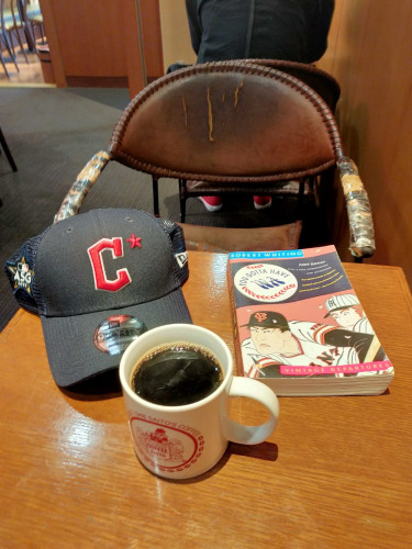 Photo is of a coffee house table. In front is a white mug of black coffee with the red medieval timey logo. Behind it to the left is a Cleveland Guardians dark blue baseball cap from the 2023 All Star Game logo on the left and a bid red C with an asterisk on the front & a sticker on the bill. To the right is the paperback book with has a baseball with the title on the upper left and Japanese  woodblock type images of Japanese men, one in a Yomiuri Giants hat & white jersey & the other has a Hanshin Tigers cap. An old tattered leather chair is in the distance. We can see a man's back & red tennis shoes can be see in the seat in front