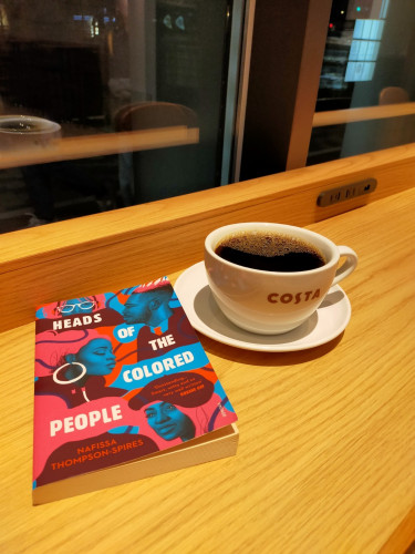 The photo is of the paperback book which is a mosaic of 3 Black women & 1 Black man in shades of blue surrounded by colors of pink & maroon. To the left on the wooden counter is a white mug & saucer of black coffee with COSTA on the mug. The window shows it is night time.