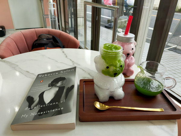 Photo is of the inside of the cafe. The table is white faux marble. The paperback book is to the left featuring a black and white photo of a white woman dressed in a 1920s outfit of black pill hat with mesh veil in front, hair tied up in a bun in the back. She looks to her right. Her black elbow length gloves are up on the wall. She has a backless V exposing her back. To the right is a darker brown rectangular tray. In front is a long gold spoon. A glass in the shape of a bear is full of shaved ice with green kiwi sauce oozing down the face. To the right on the tray is a glass pitcher of the same kiwi sauce. A plastic bear glass is behind the tray and full of strawberry milk making it look pink with a silver lid and red straw. The glass door and wall can be seen in the distance with a bicyclist pedaling past heading left