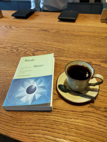 On a wooden table, at the end of which 2 flat grey scales can be seen, sits the paperback book with the top half mint green and the bottom half a blue abstract image. To the left is a mustard yellow mug and saucer of black coffee with a tiny metal spoon laying in front