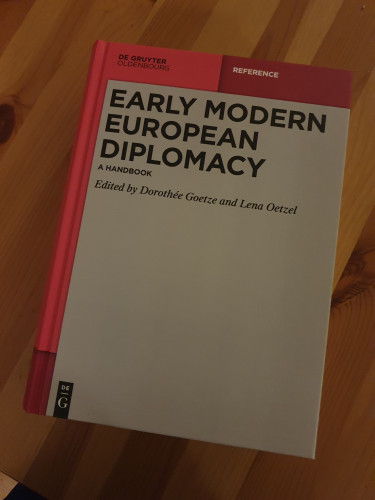 Early modern European Diplomacy. A handbook, ed. by Dorothée Goetze and Lena Oetzel. Two sorts of pink and grey cover. The book ließ on a wooden table.