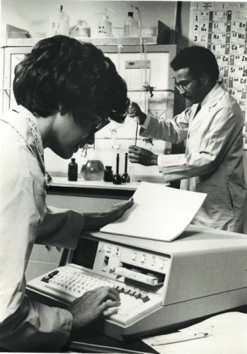 Black and white portrait orientation photograph. In the foreground is a person using the IBM 5100, one of the first portable computers, combined a typewriter-like electronic keyboard, a 10-keypad for data entry, a 1024-character display, a processing unit with up to 64K positions of main storage, and a tape cartridge for storing data. (1975). In the background is a second man with chemistry glassware equipment working with transferring liquids into test tubes.