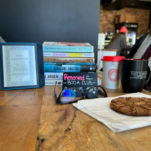 Wooden table at East Toronto Coffee Co, with "Reserved for Book Club" sign, eyeglasses, large ginger cookie, warm beverages and a stack of books, including titles by Tove Ditlevsen, Ann Patchett, Marlowe Granados, Canisia Lubrin + more