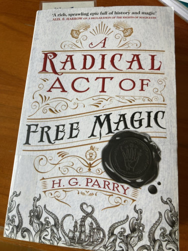 The cover of A Radical Act of Free Magic. In white with black, red & gold detail. Including a ship with a kraken wrapped around it