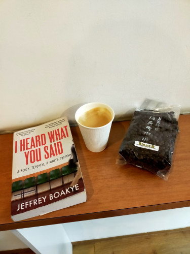 The photo is of a brown wooden counter against a white wall. The paperback book is to the left and features a photo of five green chairs and a person rushed off to the right. You can only see one khaki wearing leg and their black shoulder bag. To its right is a white paper cup of black coffee with tannish crema. To its right is a clear plastic bag of coffee beans with a lower sticker saying BLEND III and 2 vertical rows of white kanji vertical on the middle of the bag