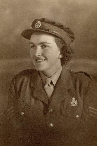 Rena Stewart in WWII 1940s ATS uniform, cap, jacket, shirt & tie. Sepia tinted black & white photo. Young white woman, dark hair, smiling looking photo left in half profile