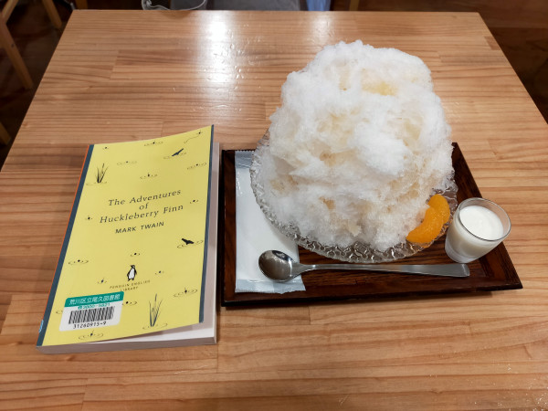 The photo is of a table in the cafe. On the left is a yellow paperback library book. On the right on is a dark wooden tray with a white mound of shaved ice with orange piece in lower right next to a tiny glass thimble of condensed milk. A long silver spoon facing left lies parallel to the tray. A plastic encased wet towel runs perpendicular on the left of the tray