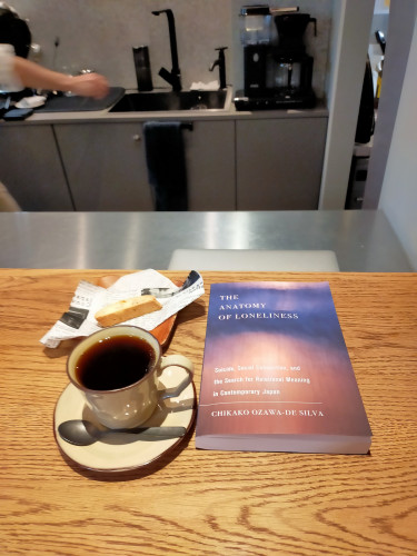 The photo is of a wooden counter in the cafe where the sink and coffee machine can be seen in the distance, as can a right arm on the left. On the counter is a light brown mug & saucer of black coffee with a silver spoon perpendicular on the saucer. The paperback book is to the left and has a Rothko blue & sienna painting cover. In the left corner is a small light brown biscotti on newspaper