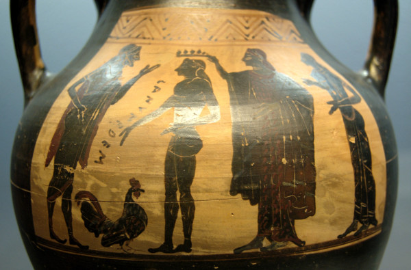 Black-figure vase painting of Zeus, Ganymedes, an unidentified goddess, and Hebe. Zeus stands to the left, one arm raised, the other gesturing towards a cock (as in rooster) on the ground. Ganymedes is facing him, also gesturing towards the animal. Both Zeus and him are in the nude, but Zeus is wearing a himation over his shoulders. Behind Ganymedes stands the unknown goddess in the process of crowning him. She is dressed in a long dress and a large himation. Behind her, Hebe watches the scene. The name "Ganymedes" is written in Greek letters between Zeus and Ganymedes.
