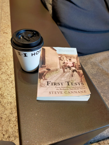 The photo is of the paperback book on a metal table with a photo of white boys playing cricket in a garage lane. The boys are all fielding with their hand on their knees wearing shorts and long sleeve jumpers. An adult white male is batting left handed in front of a box as a wicket stump. There is a red car in the distance contrasting the otherwise sepia tone of the photo. Beside the book is a white paper coffee cup with a stylish serif-ed capital H. Another bold H & O can be seen on the cup with the name cut off of Horiguchi. The cup has a black lid.