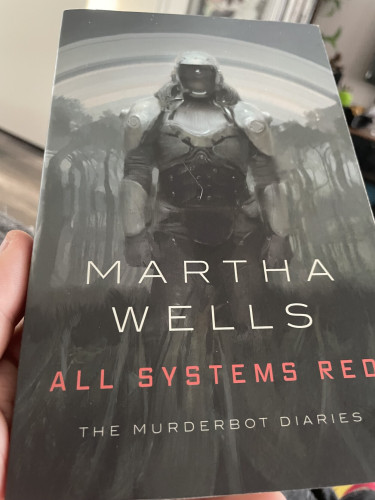 Foto van het boek All Systems Red van Martha Wells. Een hoes met veel grijstinten dat een lege planeet lijkt voor te stellen met een ruimtepak op de voorgrond. De titel valt erg op in rode letters.