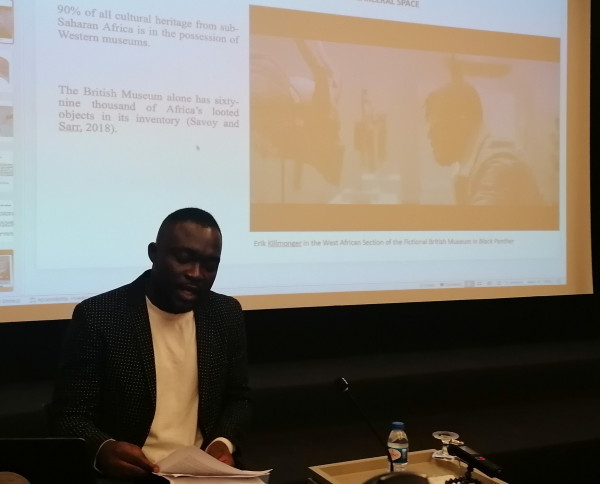 Sakiru Adebayo is sitting on the stage while giving his lecture. Behind him, the presentation showcases a frame of Blackpanther where Killmonger is looking at African art in the film's Museum. 