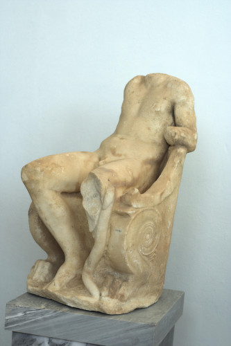 Marble sculpture of Dionysos seated on a throne in a laid back, casual way. He is missing his head, his right arm and part of his left leg but his left arm is leaning on the throne.