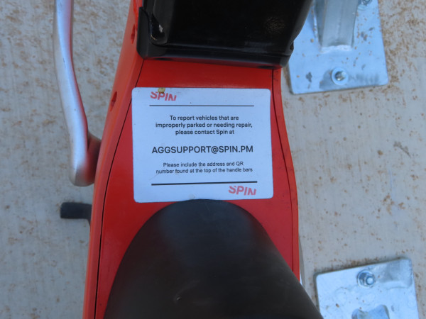 Photo of the frame of a SPIN bike.  "To report vehicles that are improperly parked or needing repair, ) ' please contact Spin at  AGGSUPPORT@SPIN.PM
Please include the address and the QR number found at the top of the handle bars."