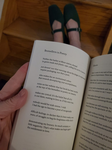 Poetry collection Stranger by Nyla Matuk (Vehicule Press), open to the poem "Bestsellers in Poetry" is held up in front of my feet in green mary janes, resting on a wooden staircase