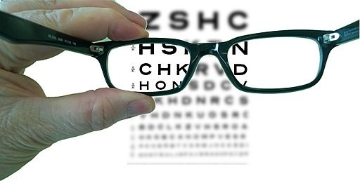 Image: holding eyeglasses in front of eye chart.