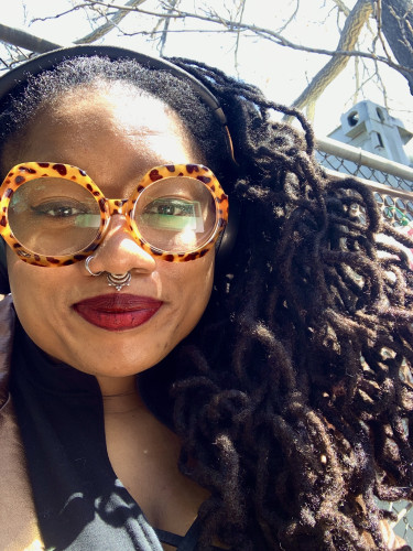 a photo of a black person sitting outside, in front of a fence and a tree. they have long curly locs that are not twisted at the root. they are wearing large round leopard print glasses, dark brown eyeliner, and deep burgundy-red lip stain. they are also wearing black and gold headphones, a black jumpsuit ,and a bronze leather jacket, which you can only see at the bottom half of the photo. they are looking directly at the camera. 