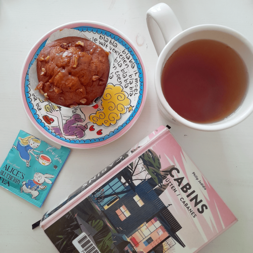 A flat lay photo of a book titles "Cabins" at the bottom. Above the book, to the right is a large cup of tea. To the left is a pecan muffin on a plate with an illustration of a blond woman with lots of text saying "blabla". All the way to the left is the packaging of a bag of Alice's Blueberry Tea.