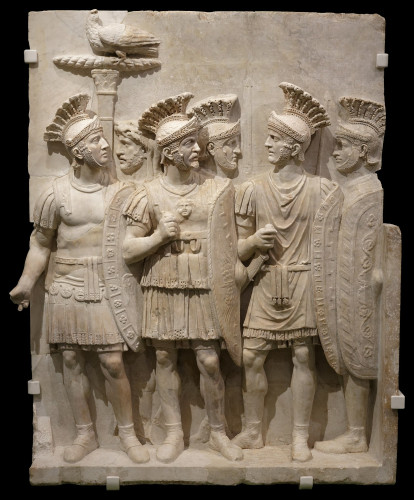 Five praetorians standing together wearing plumed helmets. Most can be seen to hold long oval shields. Another bearded male stands in the background but their identity as a praetorian is unclear.