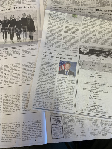Photo of a newspaper centered on an article with headline "Join Rep. Adam Kinzinger for an online program" accompanied by a color portrait of a white man in a suit in front of an American flag.