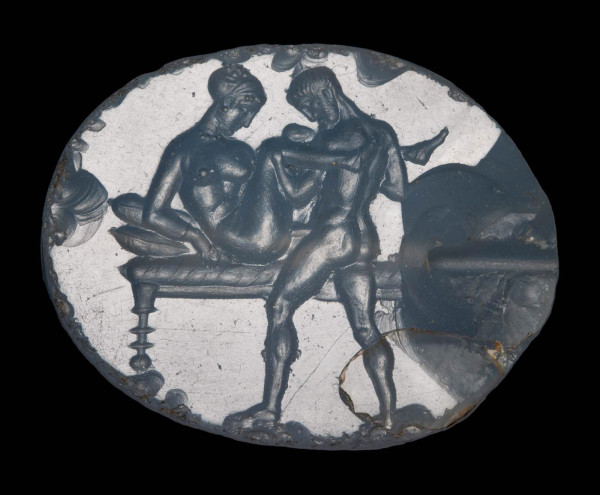 Intaglio. Pierced for stringing (horizontal). Motif oriented horizontally. A woman, seated on a cushioned bed, has intercourse with a male partner. Her face and legs are seen in profile, while her torso is twisted toward the viewer in a viewpoint somewhere between three-quarters and frontal. The woman wears a beaded necklace, a bracelet on her right arm, earrings, and thin slippers. Her hair is bound up in a scarf, with a few locks of hair escaping at her forehead and crown. She has a long nose, and heavy chin. Though seated on a bed, she holds her torso off of the bed with her arms; she may be rested her left elbow on one of the two pillows/cushions visible at the left edge of the bed. Her partner stands before the bed, and grabs her raised legs at the knees. He is bearded, and has short hair rendered by short striations. Like the woman, his face is rendered in profile, while his body is seen in three-quarter view from the back. Only one leg of the bed is visible at the left, the artist having omitted the back left leg; the right leg(s) is missing due to a large chip. Both figures have their gaze pointed down. Significant chip around right perforation.