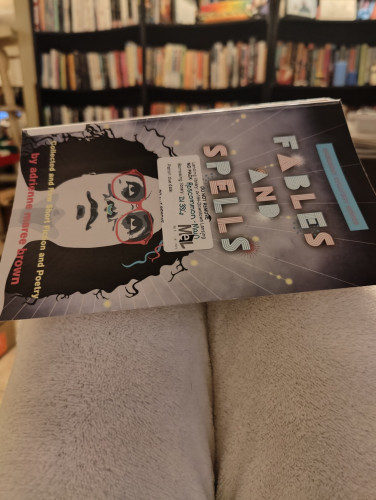 Photo of the book Fables and Spells by adrienne maree brown sitting on bent knees. Book has a picture of brown on the cover with glasses and her iconic septum ring. Bookshelves can be seen in the background.