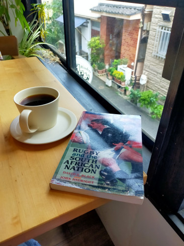 Photo is of a 2nd floor of a cafe where the alley below can be seen from the window. On the light brown table is a grey mug & saucer of black coffee. To the right is the paperback book with a close-up image of rugby players. Due to the glare, only a white player in a black New Zealand All Black jersey & white shorts holding a white rugby ball in his right hand and looking right