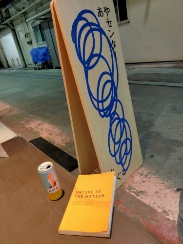 Photo is an outside bench. The orange book sits on it. To the left is a can of Gatao white wine with a cat on it  In front is a long brown sandwich board with blue scribble circles. The bench looks out in to concrete walkway underneath a train