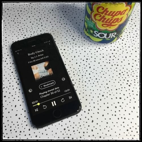 An iPhone with the cover of Body Check by J.J. Arias, narrated by Carrie Coello, on a white background with black dots in various sizes and shapes. Next to the phone, a can of Chupa Chups sour drink.