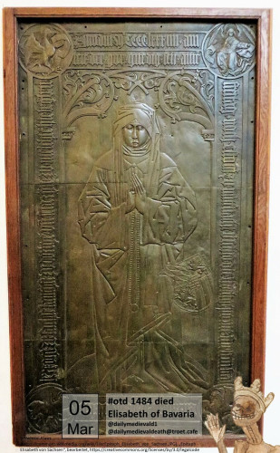 The picture shows a bronze grave slab in a wooden frame. The grave slab shows a woman with closed eyes, wearing a robe with folded hands.