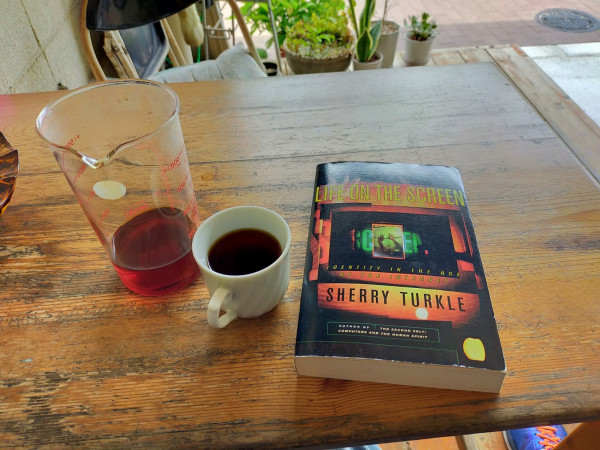 The photo is of a brown wooden table outside a coffee house. The black paperback book has an image like a movie theater but a tiny phone screen of arms raised in the center. To the left is a white mug of black coffee. Further left is a large handleless pitcher of remaining coffee