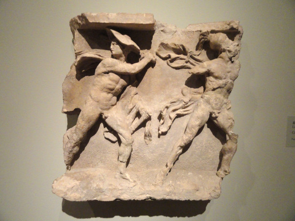 This carved limestone relief depicts two warriors striding to the right, one wearing a broad traveler's hat called a petasos and the other a Corinthian helmet. Each wears a short cloak called a chlamys over his left arm (now missing on the righthand warrior). The left warrior looks back, perhaps toward a horseman, since a small fragment of a horse’s foreleg remains behind his knee. The strong diagonals of the warriors contrasted with the flowing drapery of their cloaks demonstrates a dynamism characteristic of Hellenistic relief sculpture.