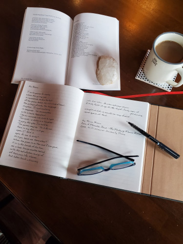 Handwritten transcription of the poem "Ars Poetica" from the poetry collection A Possible Trust by Ronna Bloom - a small piece of quartz holds open the book, an uncapped black pen and blue-framed glasses rest on the notebook page and coffee in a cream-coloured cup is nearby