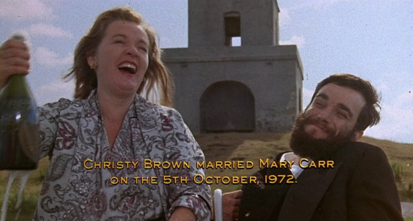 Two people, a woman who is standing and a man who is seated in a wheelchair. The man has cerebral palsy. The woman is holding a champagne bottle. Behind them there is a church on a hillside. The woman is popping the champagne and both are smiling. The caption reads, "Christy Brown married Mary Carr on the 5th October, 1972"